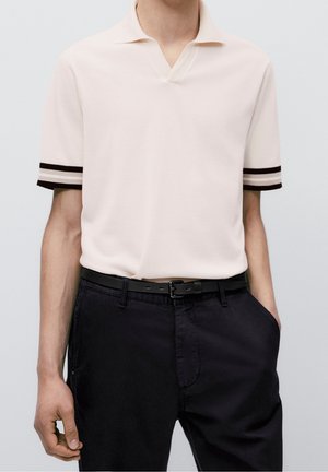 Personne portant un polo blanc à manches courtes avec des poignets rayés noir et blanc, ceinture noire et pantalon noir, une main dans la poche.