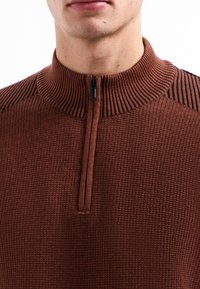 Gros plan sur une personne portant un pull en maille côtelée marron avec un col montant accentué de noir et une courte fermeture éclair à l'avant.