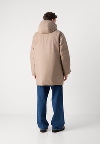 Beige péřová bunda s velkou kapucí, dlouhými rukávy a rovný střih. Nosená přes volné modré džíny, s hladkou, matnou strukturou.
