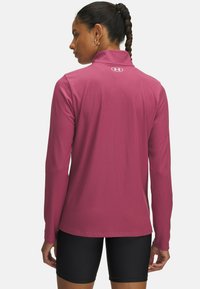 Langærmet atletisk top i et tekstureret magenta stof. Har en høj krave og Under Armour-logoet på ryggen. Stylet med sorte shorts.