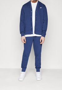 Modrý pletený cardigan s pěti knoflíky, nošený přes bílé tričko a doplněný modrými tepláky. Outfit doplňují bílé tenisky.