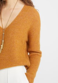 Musztardowy żółty sweter z prążkowanego materiału, z dekoltem w kształcie litery V i długimi rękawami, w połączeniu z białym dołem o wysokim stanie i naszyjnikiem z złotymi akcentami.