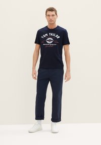 Marineblaue Baumwoll-T-Shirt mit weißem und rotem Text; kombiniert mit dunkelblauen Hosen; Modell trägt weiße Turnschuhe und Knöchel-Socken.