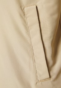 Texture di tessuto beige con un semplice dettaglio di tasca stretta. Il materiale appare liscio con una lucentezza minima, con cuciture pulite lungo i bordi.
