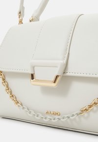 Sac à main en simili cuir blanc avec chaîne dorée et quincaillerie d'accent, doté d'un rabat et d'un design structuré. Logo de la marque visible.