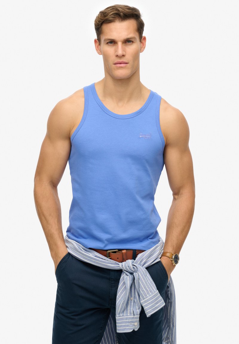Top de tirantes azul de algodón con un escote redondo, que presenta un logo bordado en el pecho. Combinado con pantalones marinos y una camisa a rayas atada a la cintura.