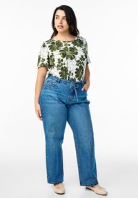 Balta marškinėliai su žaliu lapų spausdiniu, derinami su mėlynomis plačiomis džinsomis ir smėlio spalvos balerinos. Modelis stovi su rankomis kišenėse.