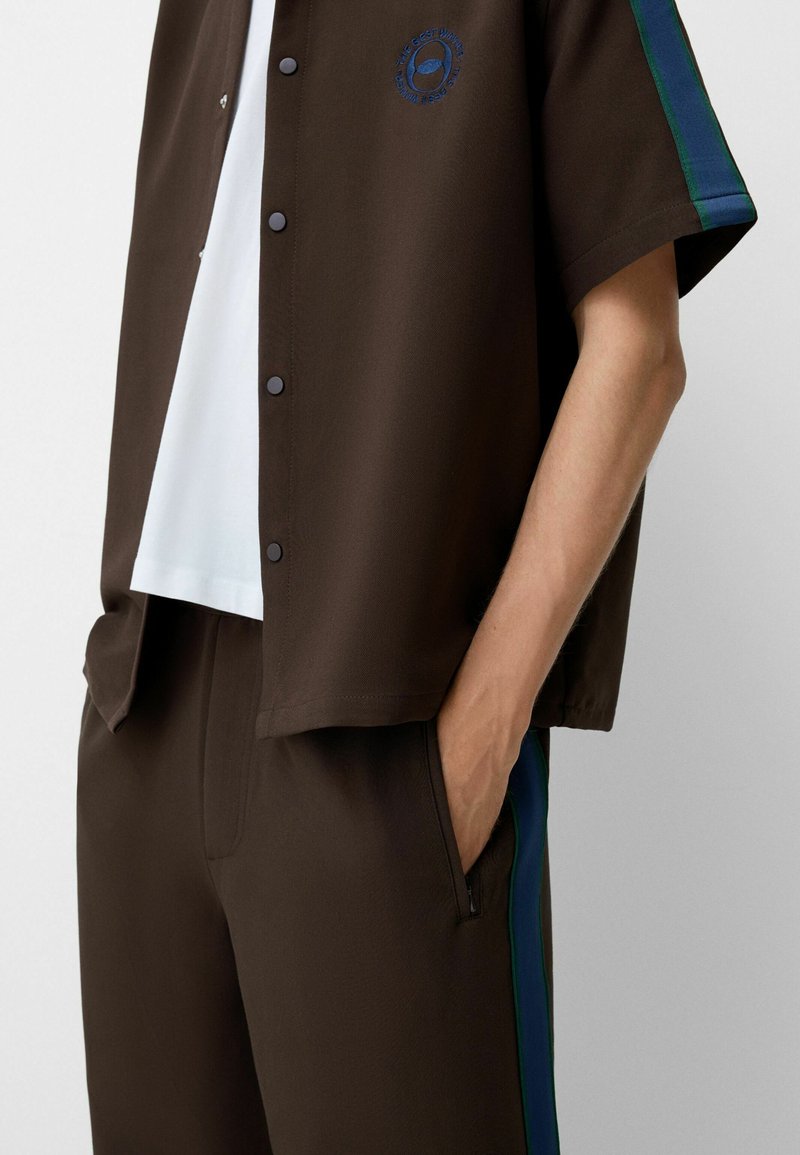 Chemise marron à manches courtes avec fermeture à bouton, assortie à un pantalon marron avec des rayures latérales bleu-vert et un détail de poche.