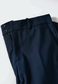 Pantalon bleu marine avec une coupe ajustée, devant plat, fermeture éclair latérale et deux petits accents métalliques près de la taille. Texture de tissu lisse.