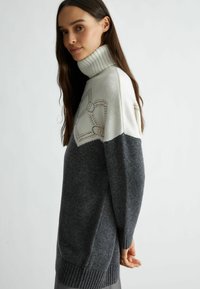 Maglione a collo alto con la parte superiore bianca e il corpo grigio scuro, caratterizzato da cuciture decorative arancioni. Texture a maglia, polsini e orlo a coste.