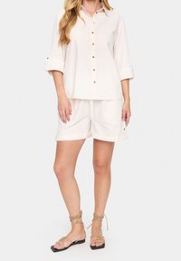 Camicia button-up beige chiaro con maniche a tre quarti, abbinata a pantaloni corti coordinati. Entrambi hanno una consistenza morbida e bordi leggermente arrotolati.