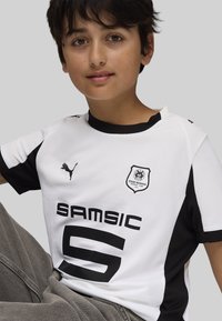 Joven vestido con una camiseta de fútbol blanca y negra del Stade Rennais, sentado con una rodilla levantada contra un fondo gris liso.