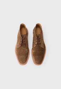 Zapatos de ante marrón con una textura suave, que presentan una punta redondeada, dos ojetes para los cordones y una suela cosida en contraste.
