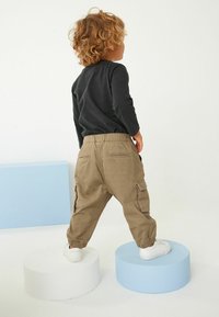 Pantalons cargo marron avec des poignets élastiques et des poches, associés à un tee-shirt noir à manches longues. L'enfant se tient sur des piédestaux ronds. Baskets blanches.