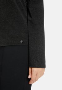 Schwarzes Langarmshirt aus strukturiertem Stoff mit einem kleinen metallischen Akzent am Saum. Der Hintergrund ist in einer dunklen einheitlichen Farbe.