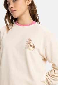 Beige sweatshirt met een ronde hals, voorzien van een bloemenprint en "ROXY" in gouden letters op de borst. Zacht, gebreid materiaal.