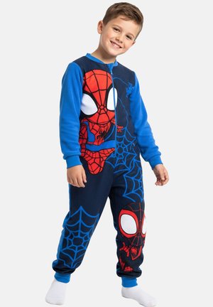 Smiling boy in blue Spider-Man onesie with web patterns and large red Spider-Man faces stands against plain background.