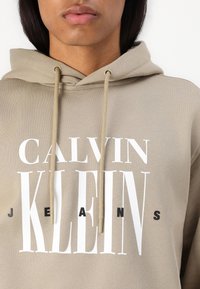 Sudadera beige con capucha ajustable que presenta en bold el texto blanco "CALVIN KLEIN" y "JEANS" en negro. Textura suave con un ajuste relajado.