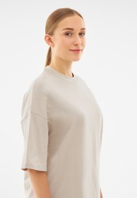 Tricou din bumbac bej deschis, cu mâneci scurte, decolteu rotund și o croială lejeră. Textură netedă, cu cusături minime și fără modele vizibile.