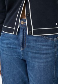 Marineblå cardigan med hvid kant, knaplukning, over blå denimjeans med en synlig kobberknap, der har en vasket tekstur og let falming.