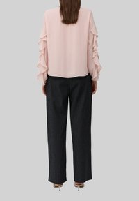 Sieviete ar gaiši rozā blūzi ar kroku piedurknēm, melnām svītrainām plašām biksēm un baltām kaķēnu papēžiem, stāv ar muguru pret skatītāju.