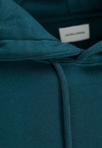 Hettegenser i dyp teal-stoff. Har en teksturert overflate og flettet snor. Merke synlig inne i kragen.