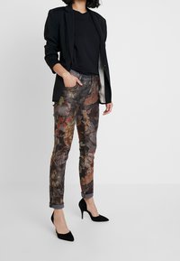 Pantaloni a stampa floreale in tonalità scure con vestibilità slim, orli risvoltati e cuciture decorative, abbinati a un blazer nero e scarpe con tacco.