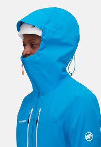 Blaue wasserdichte Jacke mit hohen Kragen, verstellbarem Kapuze und kontrastierenden weißen Reißverschlüssen. Mit einem Logo auf dem Ärmel und elastischen Kordelzügen.