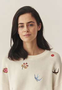 Kremowy sweter z dzianiny z kolorowym haftem kwiatów i ptaków. Posiada prążkowany dekolt i luźny krój. Teksturowana tkanina.