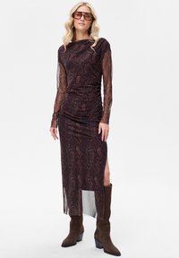 Brązowa sukienka z długim rękawem w nadruk w wężowy wzór, z marszczeniami, rozcięciem z boku i przezroczystą tkaniną. Połączona z brązowymi wysokimi kozakami za kolano.