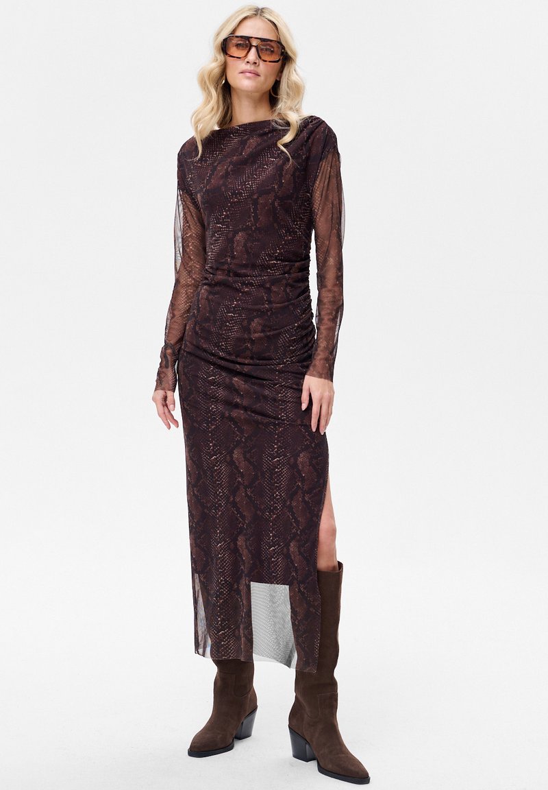 Brązowa sukienka z długim rękawem w nadruk w wężowy wzór, z marszczeniami, rozcięciem z boku i przezroczystą tkaniną. Połączona z brązowymi wysokimi kozakami za kolano.