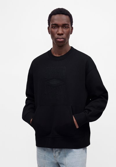 Νέος άνδρας που φοράει μαύρο φούτερ Emporio Armani με τα χέρια στην μπροστινή τσέπη, στέκεται μπροστά σε ένα απλό λευκό φόντο.