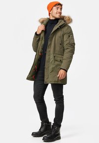 Parka de color verde oliva con capucha con borde de piel, cierre de cremallera y botones, bolsillos frontales y forro interior con un patrón rojo. Jeans negros y botas.