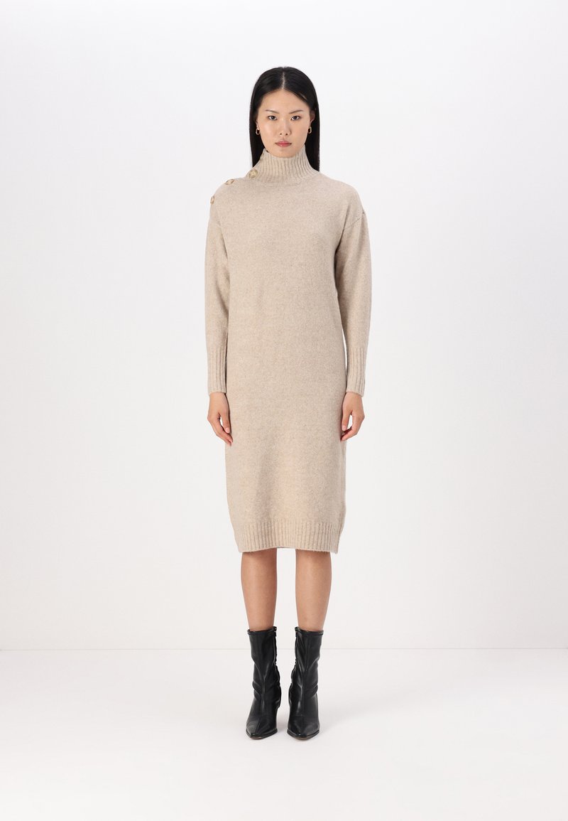Abito beige a maglia con collo alto, polsini a costine e dettagli di bottoni laterali. Il modello lo abbina a stivali neri alla caviglia. Silhouette semplice e dritta.