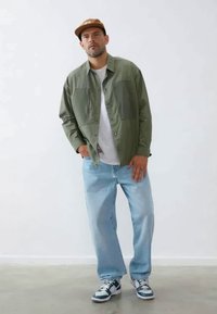 Olivgrüne Button-up-Hemd mit zwei Brusttaschen, weißem T-Shirt darunter, hellblauen Jeans und schwarz-weißen Sneakers.