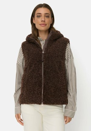 Kvinde iført en brun, lynlåsåben, fluffy vest over en beige og hvidstribet langærmet skjorte og hvide bukser mod en ensfarvet baggrund.