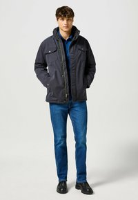 Chaqueta aislante negra con múltiples bolsillos en el pecho, un collar alto y una cremallera. Usada sobre una blusa azul, combinada con jeans azules y zapatos negros.
