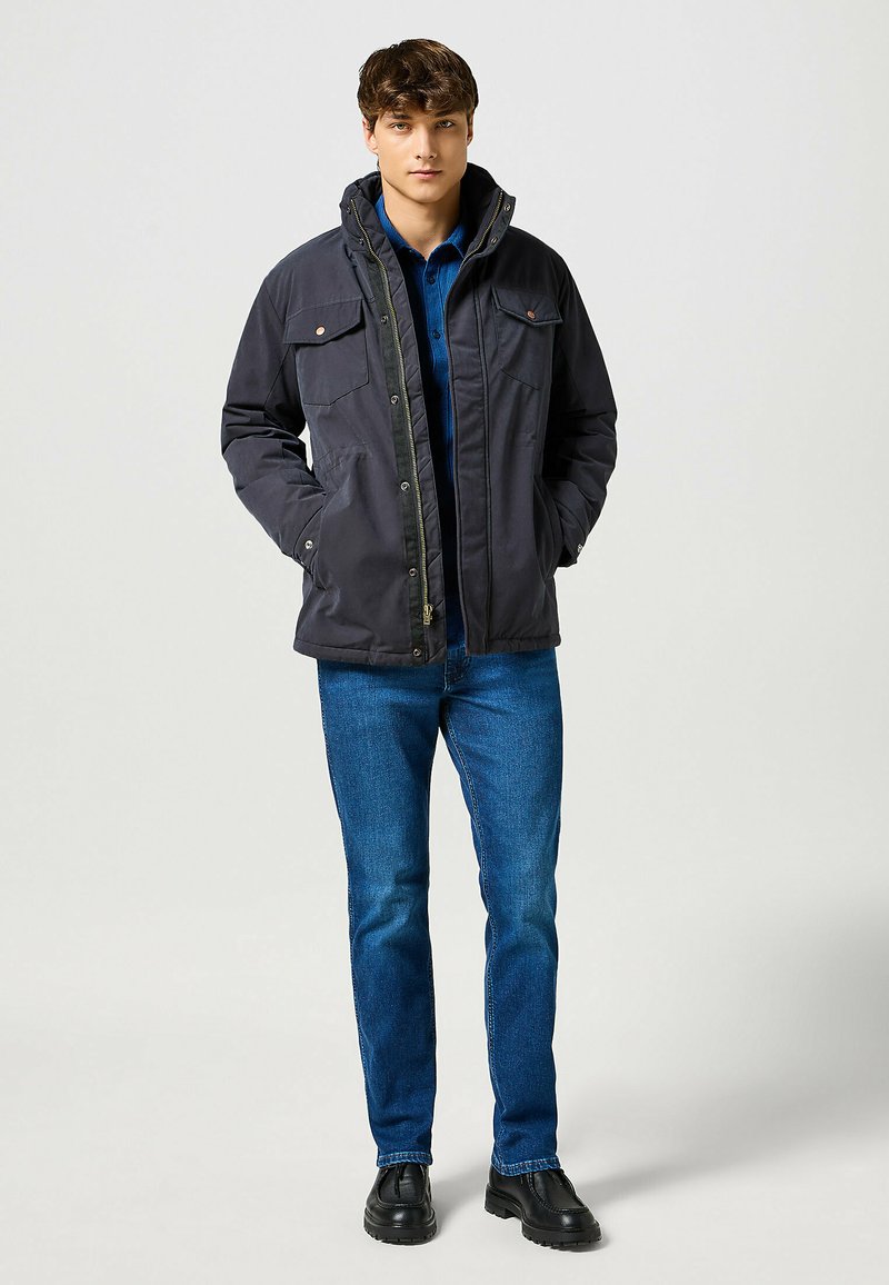 Giubbotto isolante nero con multiple tasche sul petto, colletto alto e zip. Indossato sopra una maglietta blu, abbinato a jeans blu e scarpe nere.