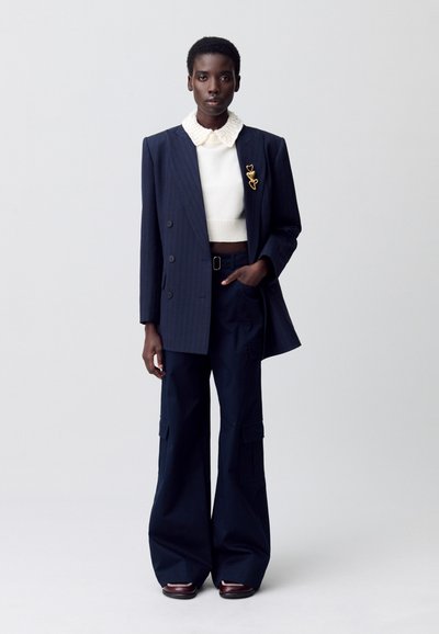 Blazer de lana con rayas marinas de doble botonadura, suéter de punto blanco, pantalones cargo de cintura alta; detalle de broche dorado, zapatos planos.