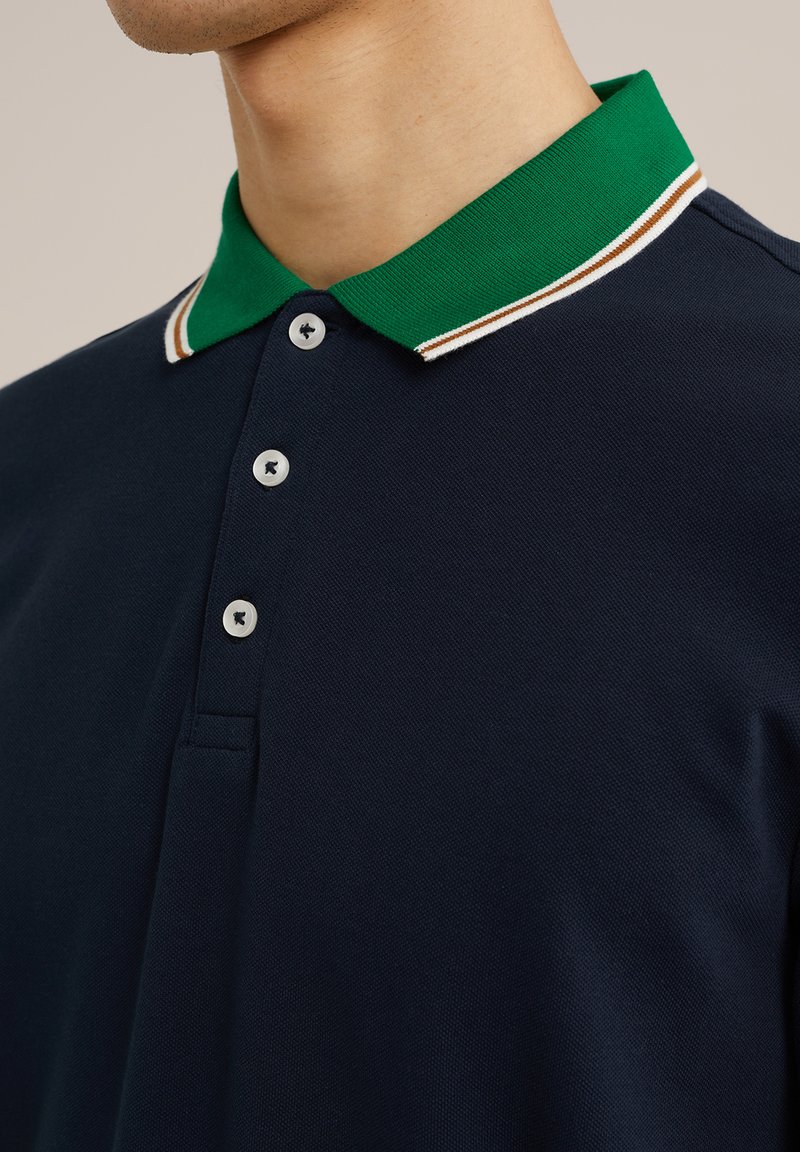Polo shirt bleu marine avec un col tricoté vert comportant des rayures blanches et marron, patte de boutonnage à trois boutons et tissu texturé.