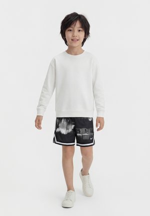 Garçon marchant portant un sweat-shirt blanc à manches longues, un short à motif abstrait noir et blanc, et des baskets blanches sur un fond uni.