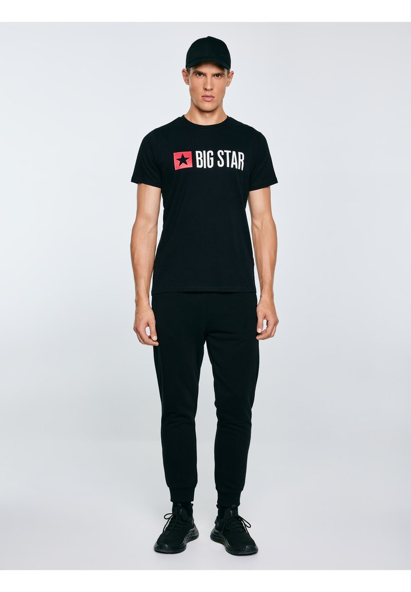 Mężczyzna w czarnej czapce, czarnej koszulce z napisem "BIG STAR", czarnych spodniach dresowych i czarnych tenisówkach, stojący na jasnym, jednolitym tle.