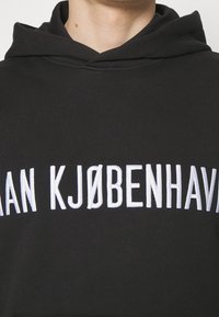 Felpa nera con cappuccio caratterizzata da testo ricamato bianco che reca la scritta "AN KJØBENHAVN". Il materiale è morbido e con una texture classica, dotata di cappuccio regolabile.