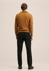 Brauner gestrickter Pullover mit rippenstruktur, kombiniert mit schwarzen Hosen. Das Ensemble zeichnet sich durch eine moderne, taillierte Silhouette und flache Schuhe aus.