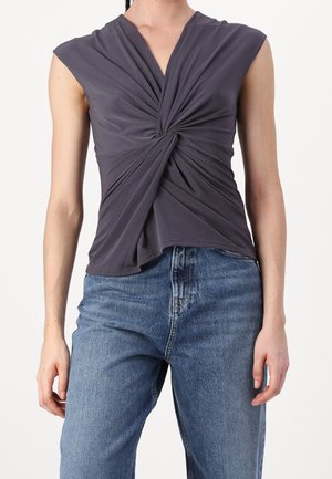Haut sans manches gris foncé avec un détail torsadé sur le devant, porté avec un jean bleu taille haute sur une personne debout sur fond blanc.