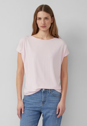 Vrouw met lang lichtbruin haar, gekleed in een lichtroze top met korte mouwen en een blauwe spijkerbroek, staand tegen een effen grijze achtergrond.