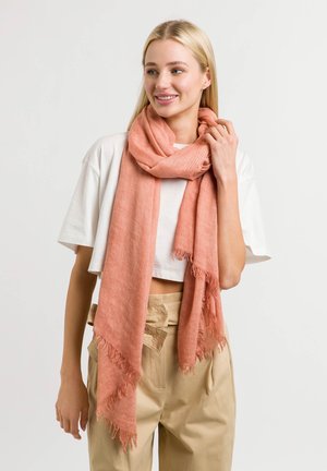 Sciarpa di coral leggero con bordi sfilacciati, indossata attorno al collo, abbinata a una camicia bianca corta e pantaloni beige, su uno sfondo neutro.
