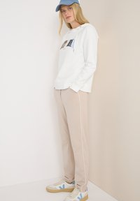 Felpa bianca con design grafico, pantaloni beige con bordino bianco e sneakers bianche con dettagli blu, abbinati a un cappello azzurro chiaro.