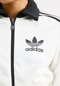 White zip-up jacket with black collar and three black stripes on shoulders. Features black adidas logo on the left chest. Smooth texture.