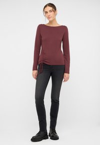 Bordowy top z długim rękawem i dekoltem łódkowym, z marszczeniem po boku, w zestawieniu z czarnymi rurkami i czarnymi butami sznurowanymi.